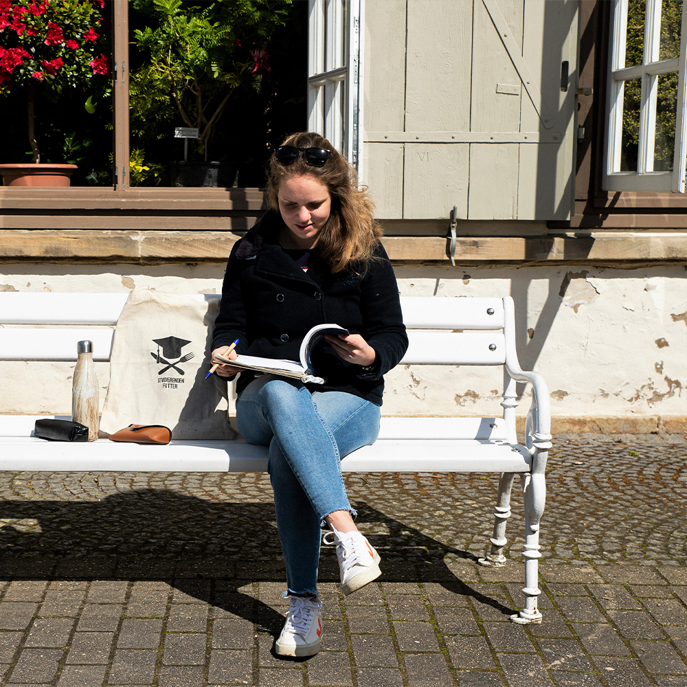 Studentin sitzt mit Lehrbuch auf einer Parkbank und Studierendenjutebeutel neben sich