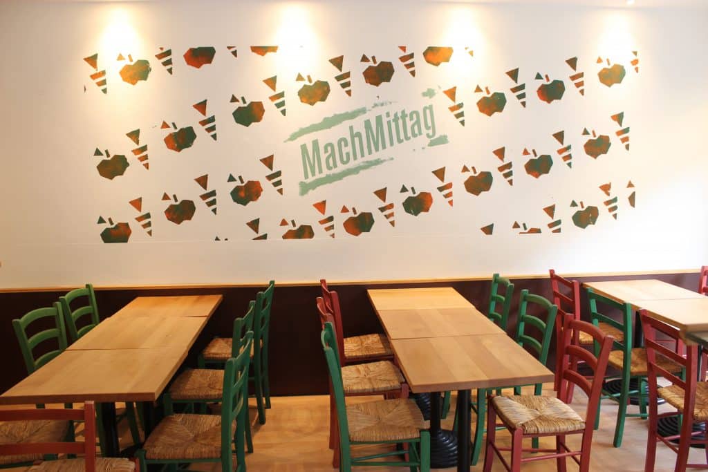 Blick in das Restaurant von Mach Mittag mit Tischen und Stühlen und einem Mach Mittag Schriftzug an der Wand.